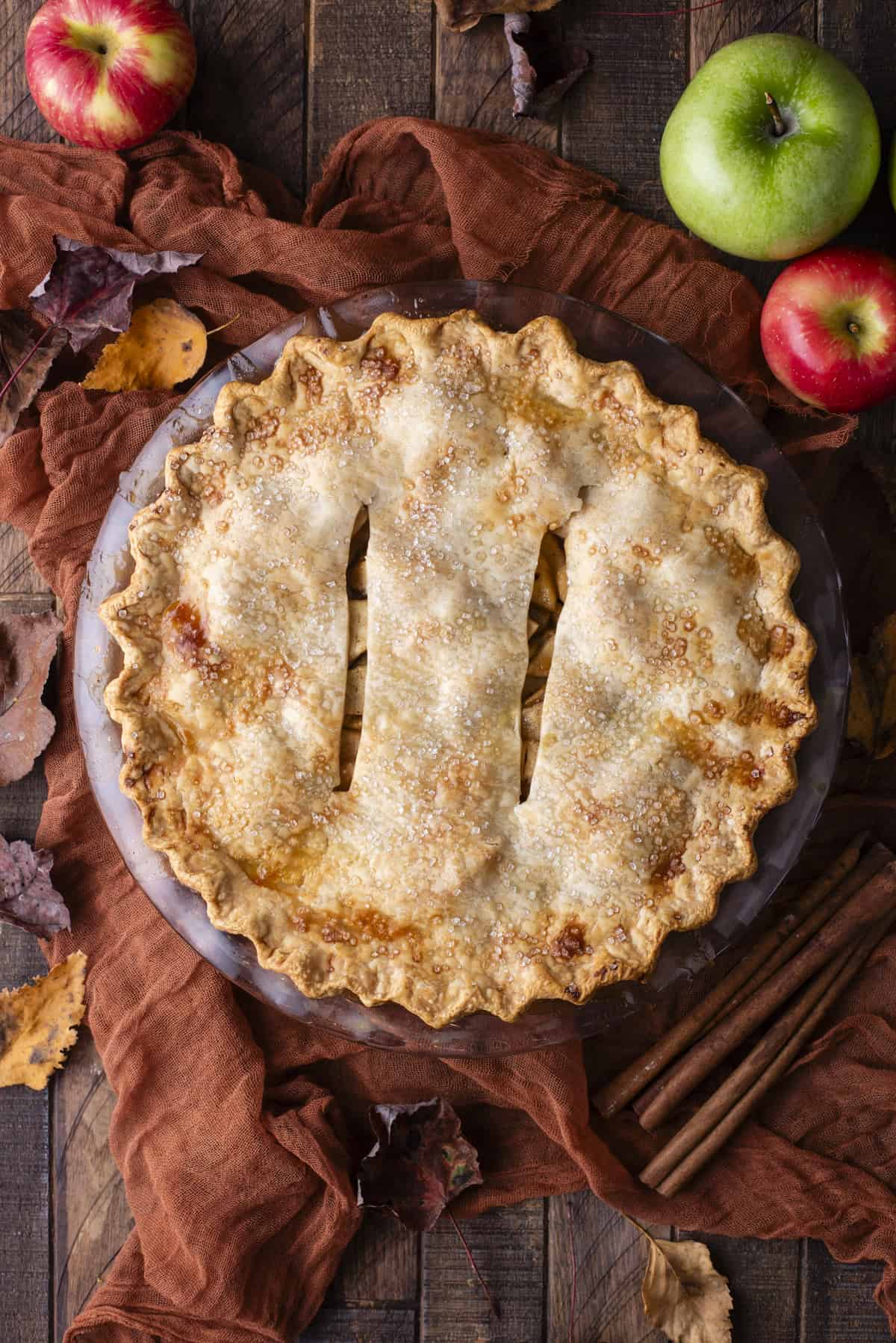 over head view of an apple pie surrounded by green and red apples, fall leaves and cinnamon sticks