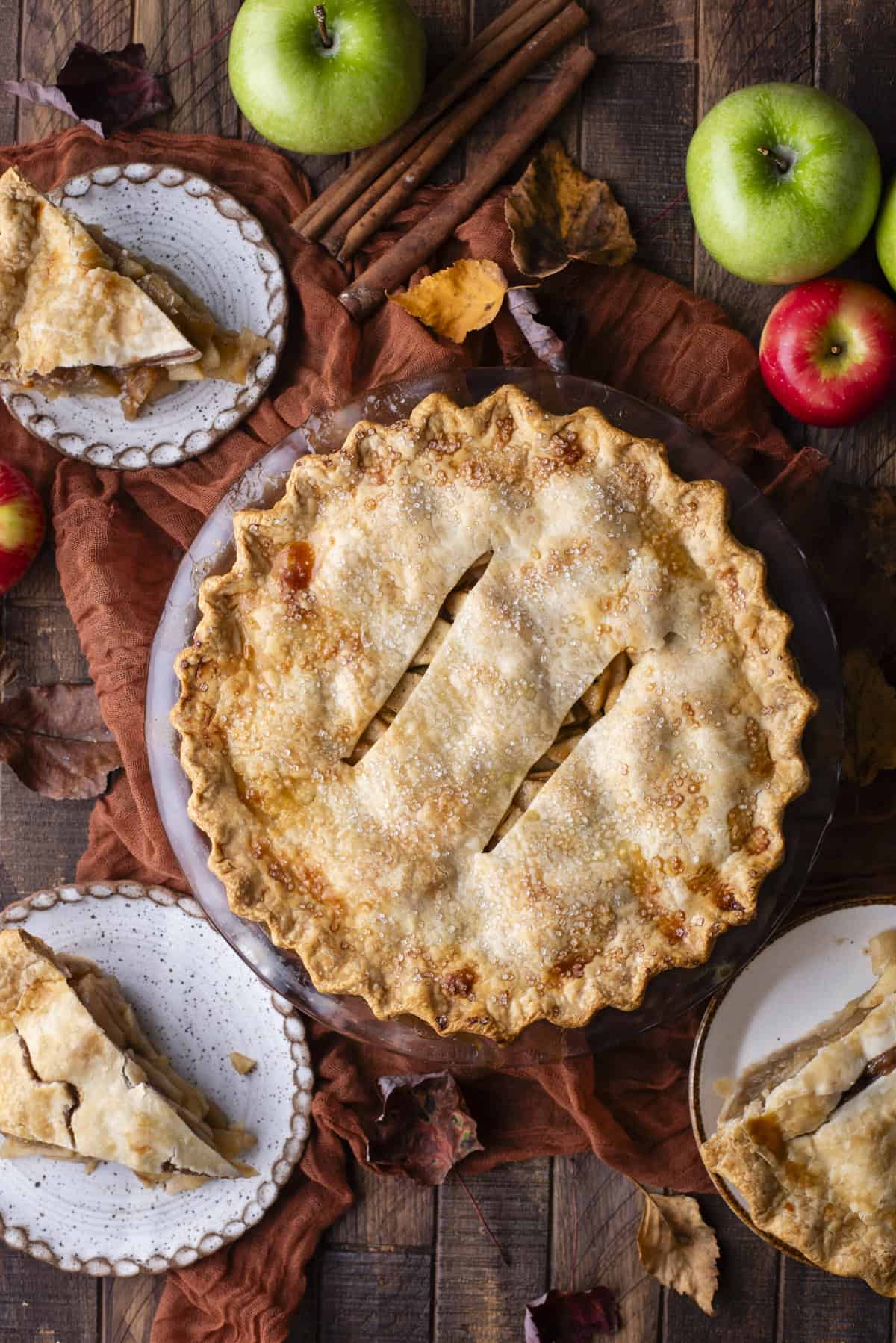 over head view of an apple pie surrounded by slices of apple pie on plates, green and red apples and fall leaves