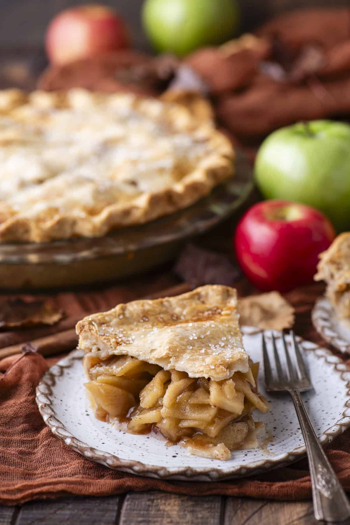 a slice of apple pie on a small plate with a fork, surrounded by red and green apples and a full pie in the background