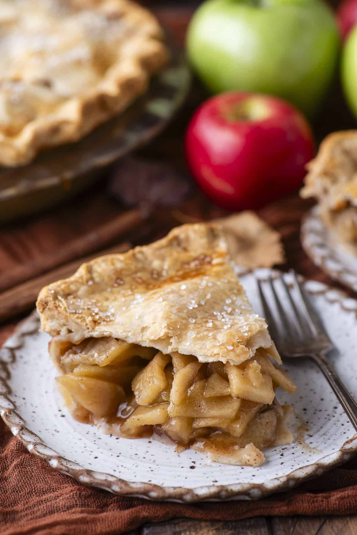 a slice of apple pie on a small plate with a fork, with red and green apples and more pie in the background