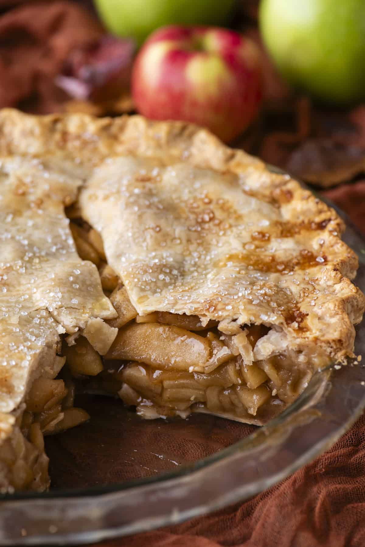 apple pie in a pie dish with one slice missing, exposing the apple filling inside, red and green apples in the background