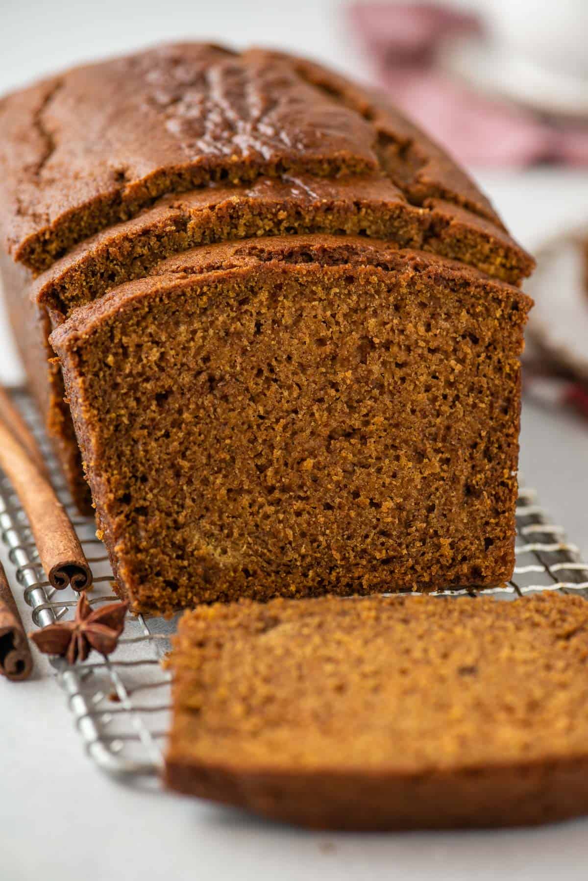 a loaf of pumpkin bread on a wire rack, with a slice cut laying by the loaf and two more slices leaning on the rest of the loaf, a cinnamon stick and star anise scattered around