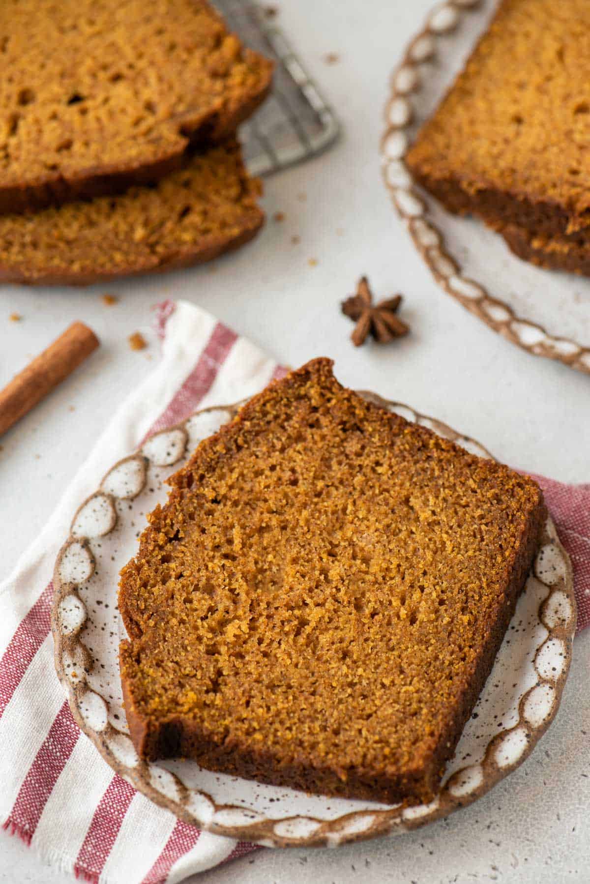 a slice of pumpkin bread on a small plate, sitting on top of a red and white striped towel with a cinnamon stick, star anise and more slices of bread surrounding it