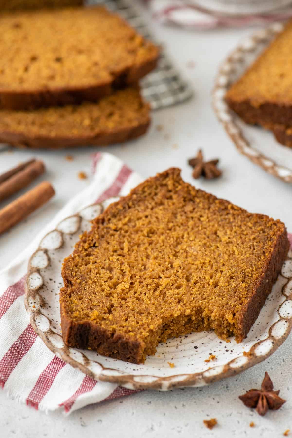 a slice of pumpkin bread on a small plate with a bite missing, sitting on top of a red and white striped towel with a cinnamon stick, star anise and more slices of bread surrounding it