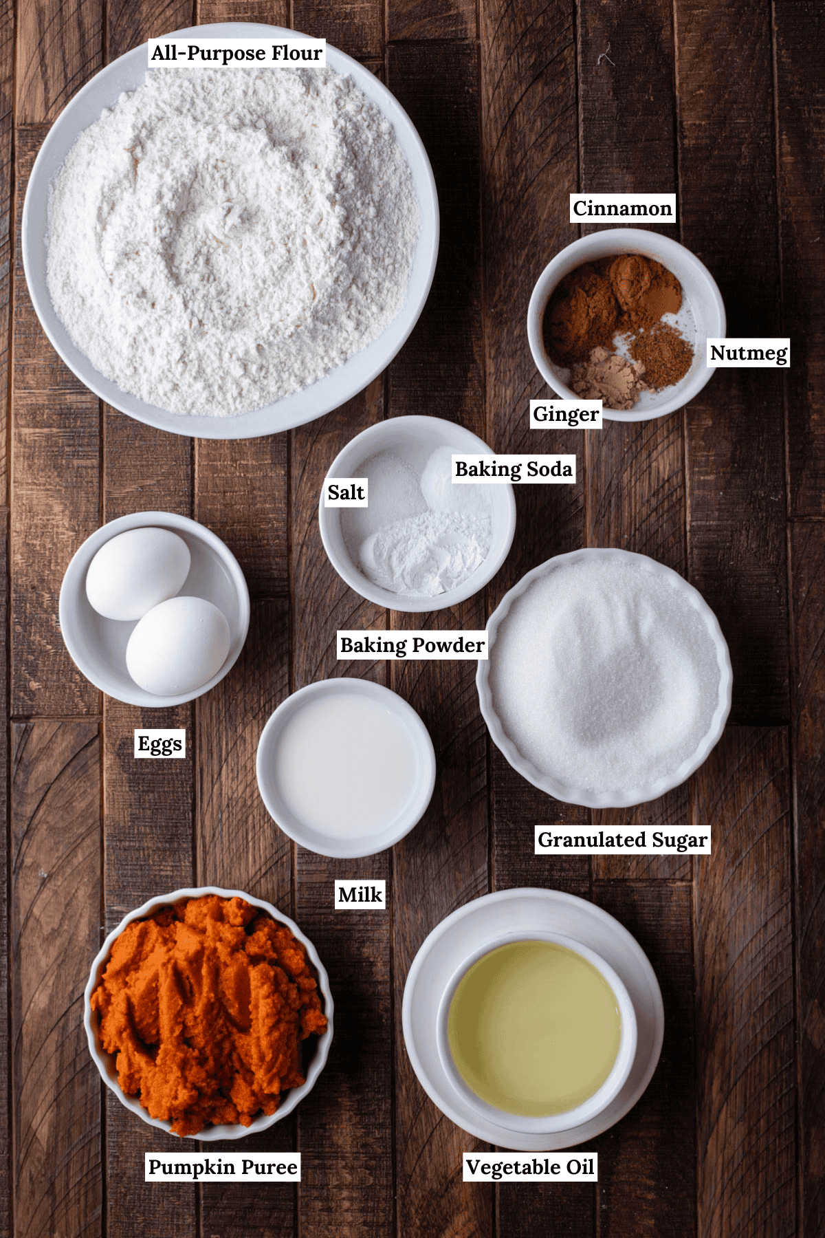 over head view of the ingredients for pumpkin bread including all-purpose flour, cinnamon, nutmeg, ginger, salt, baking soda, eggs, baking powder, granulated sugar, milk, pumpkin puree and vegetable oil