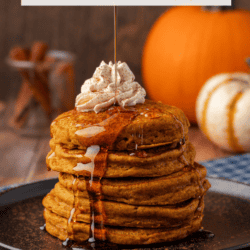 a stack of pumpkin pancakes on a black plate, topped with whipped cream, being drizzled with syrup