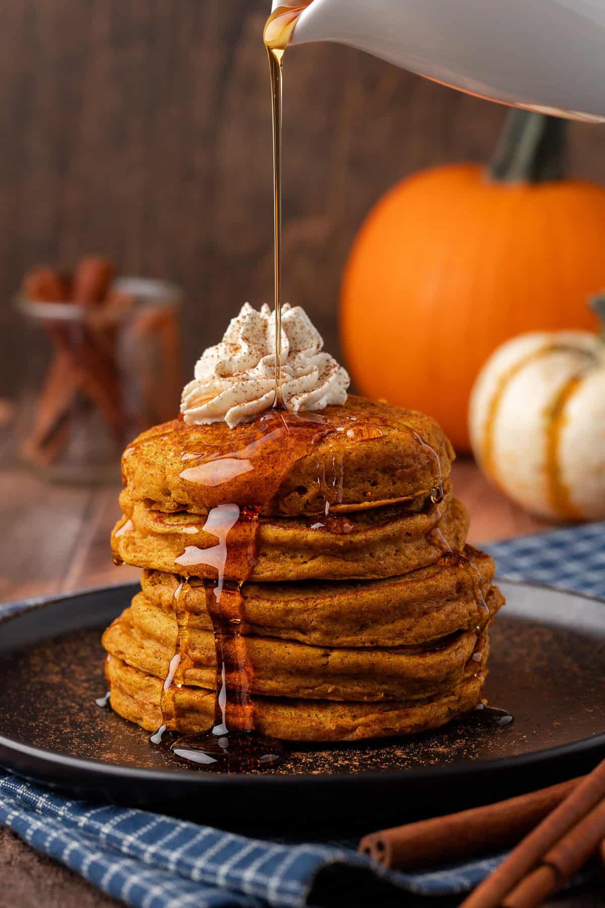 a stack of pumpkin pancakes on a black plate, topped with whipped cream, being drizzled with syrup