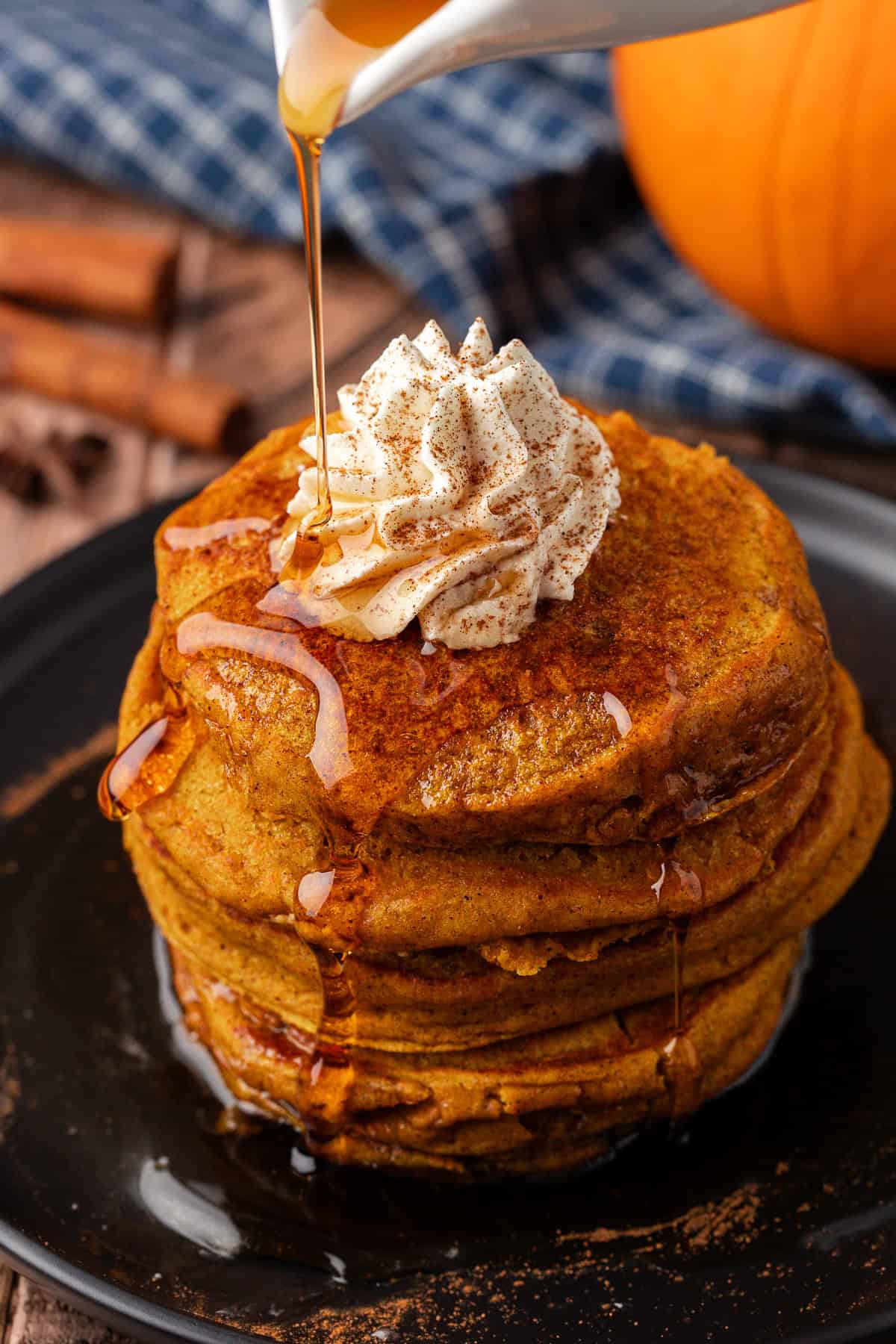 a stack of pumpkin pancakes on a black plate, topped with whipped cream, being drizzled with syrup