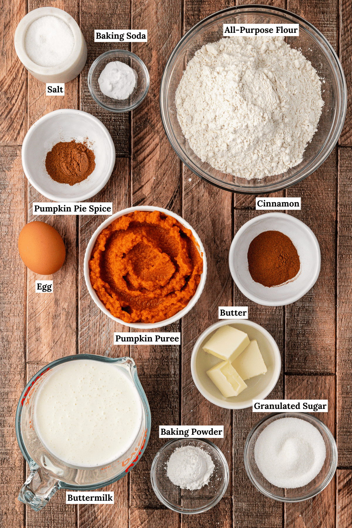 over head view of the ingredients for pumpkin pancakes including salt, baking soda, all-purpose flour, pumpkin pie spice, egg, pumpkin puree, cinnamon, butter, granulated sugar, buttermilk and baking powder