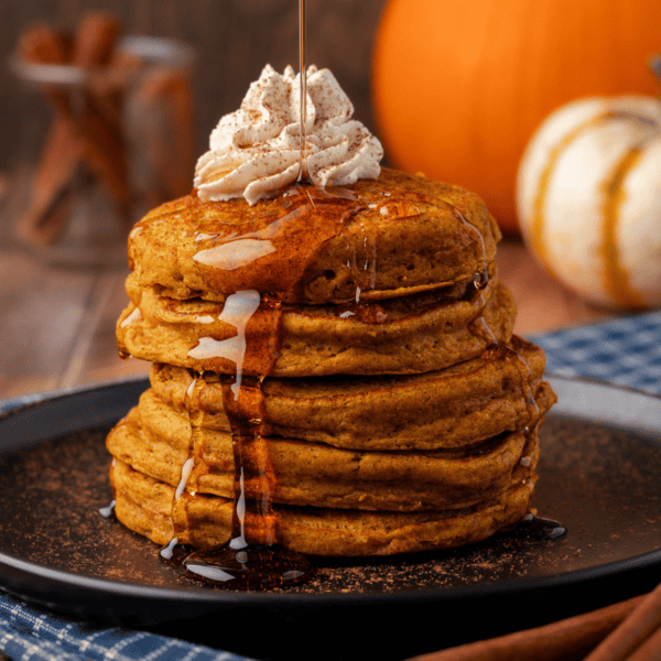 a stack of pumpkin pancakes on a black plate, topped with whipped cream, being drizzled with syrup