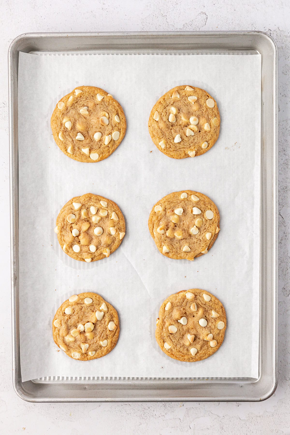 overhead view of two rows of three white chocolate chips cookies on a baking sheet lined with white parchment paper