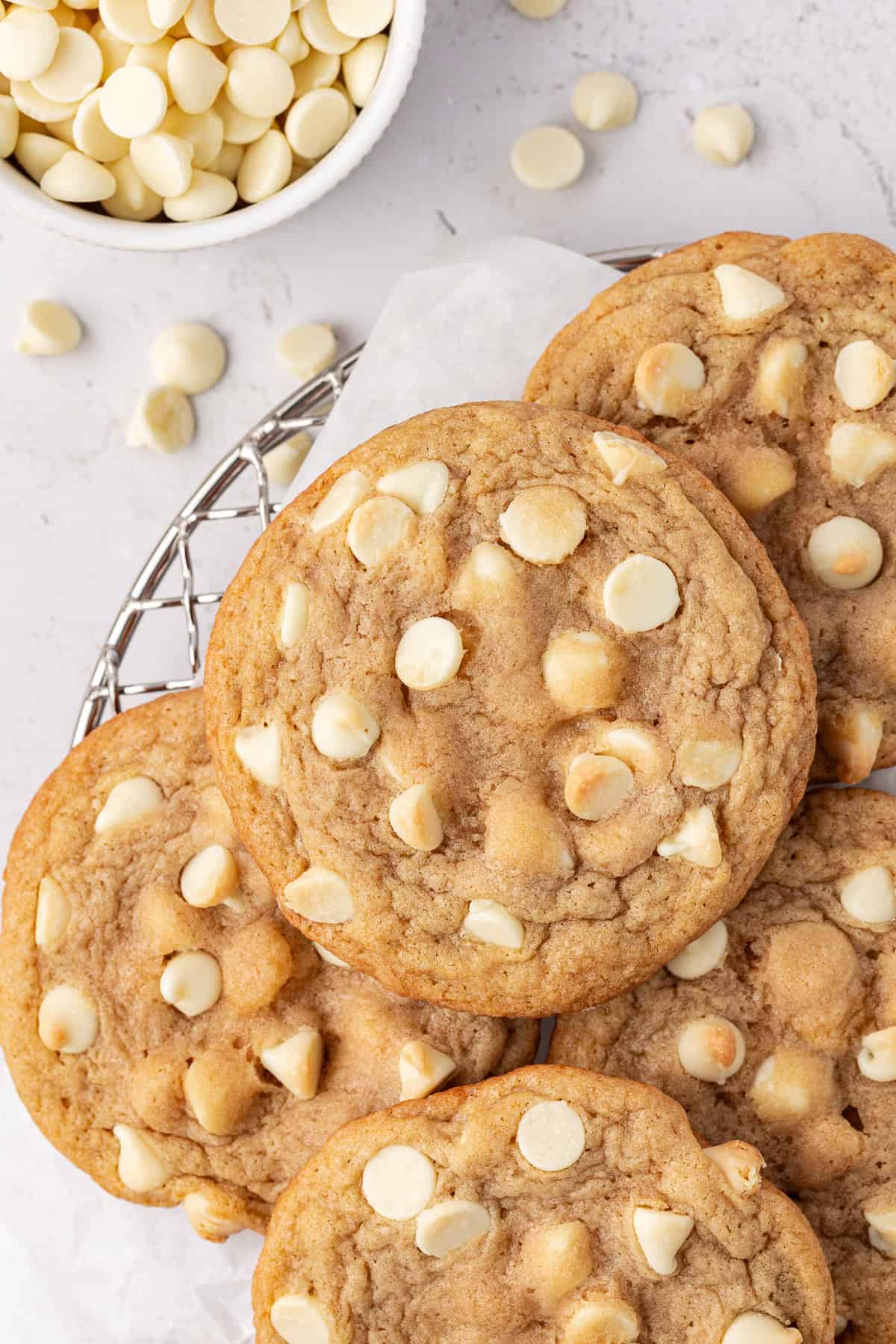 overhead view of white chocolate chip cookies piled on a wire rack with white chocolate chips in a small white bowl beside it and scattered around