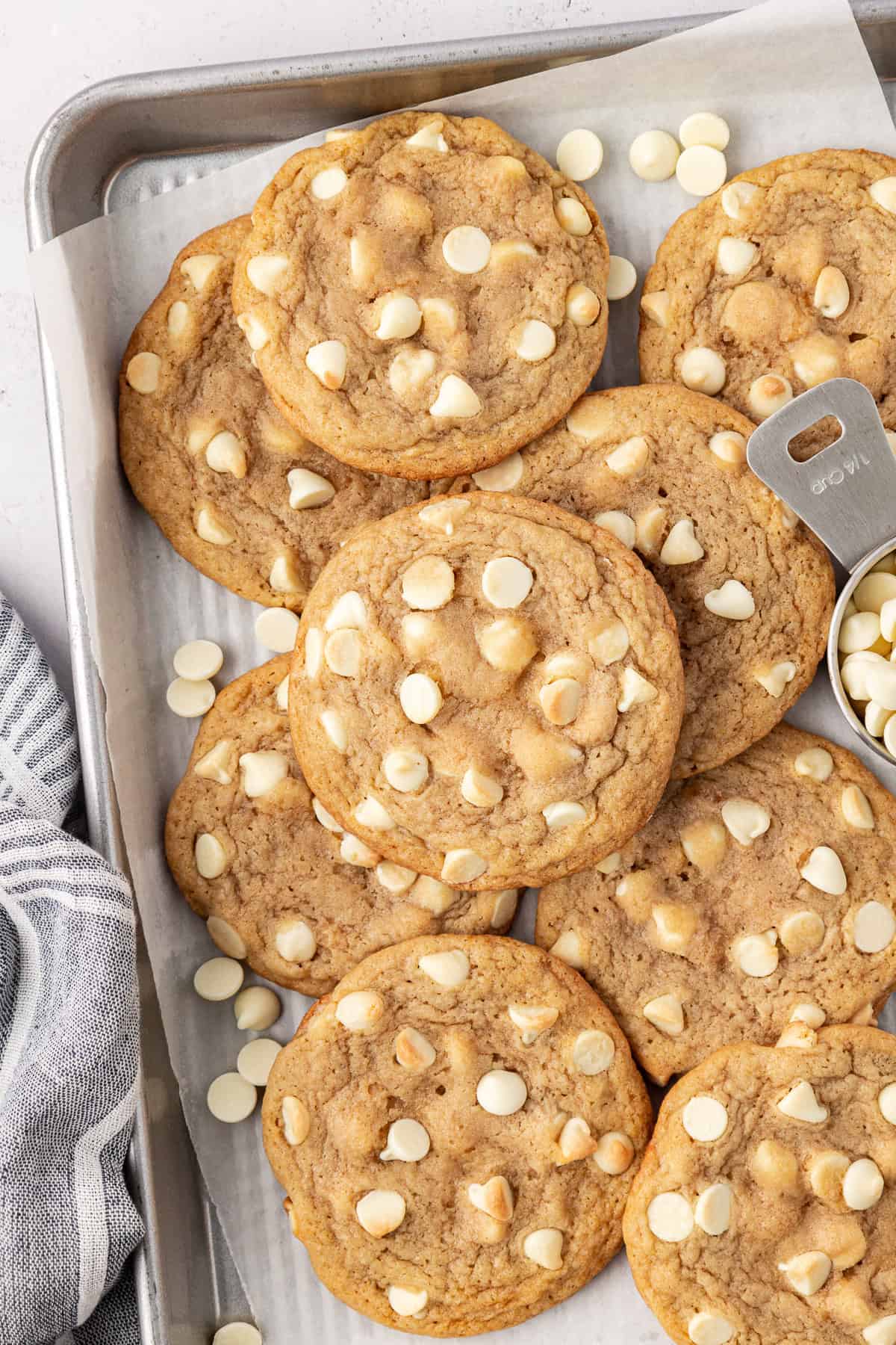 white chocolate chip cookies and white chocolate chips piled and scattered on a baking sheet lined with white parchment paper