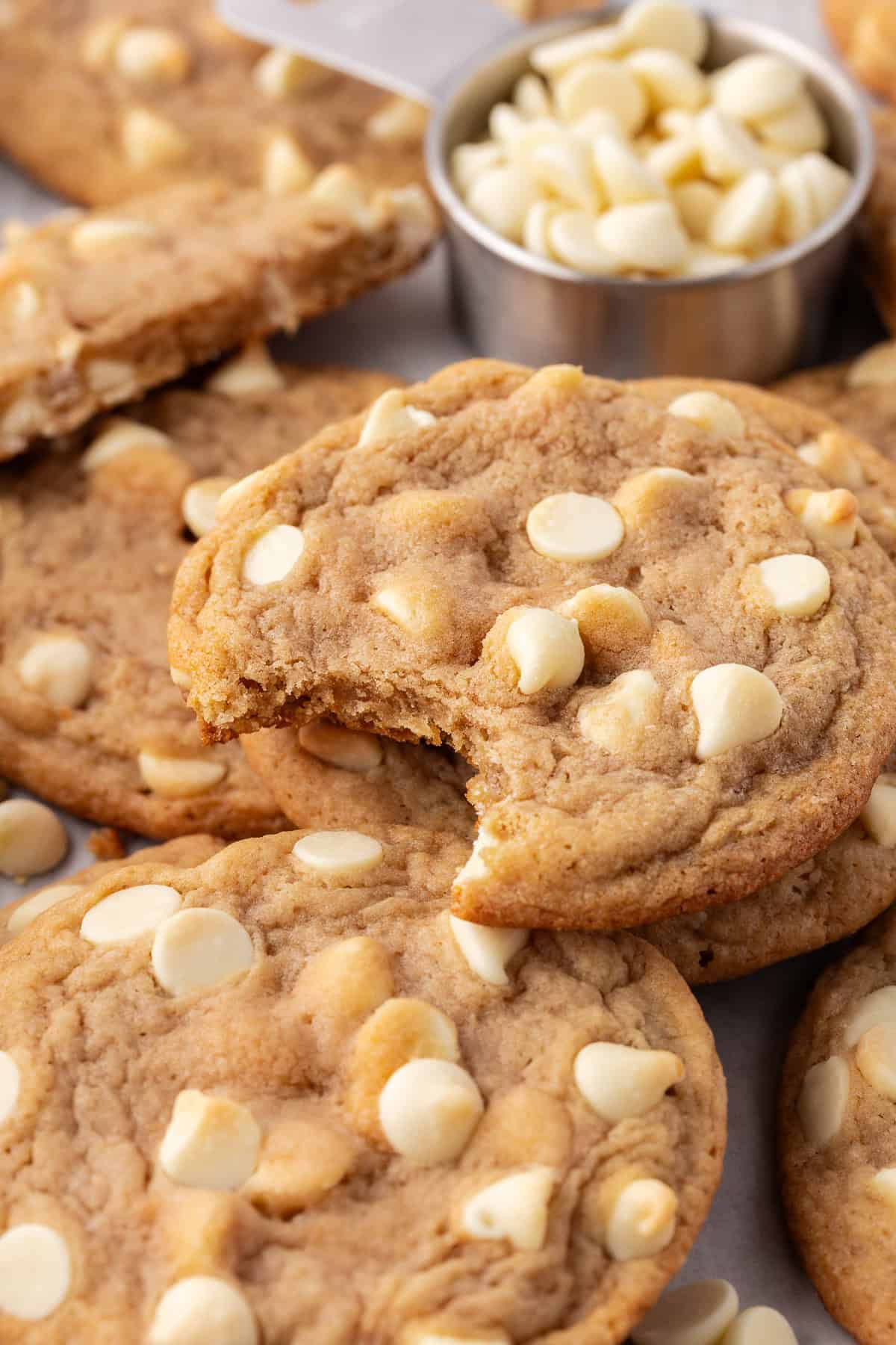 a pile of white chocolate chip cookies with a measuring cup of white chocolate chips, and a bite missing out of the top cookie