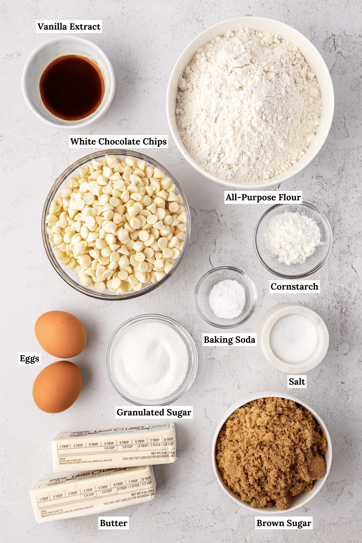 over head view of white chocolate chip cookie ingredients including vanilla extract, all-purpose flour, white chocolate chips, cornstarch, eggs, baking soda, granulated sugar, salt, butter and brown sugar