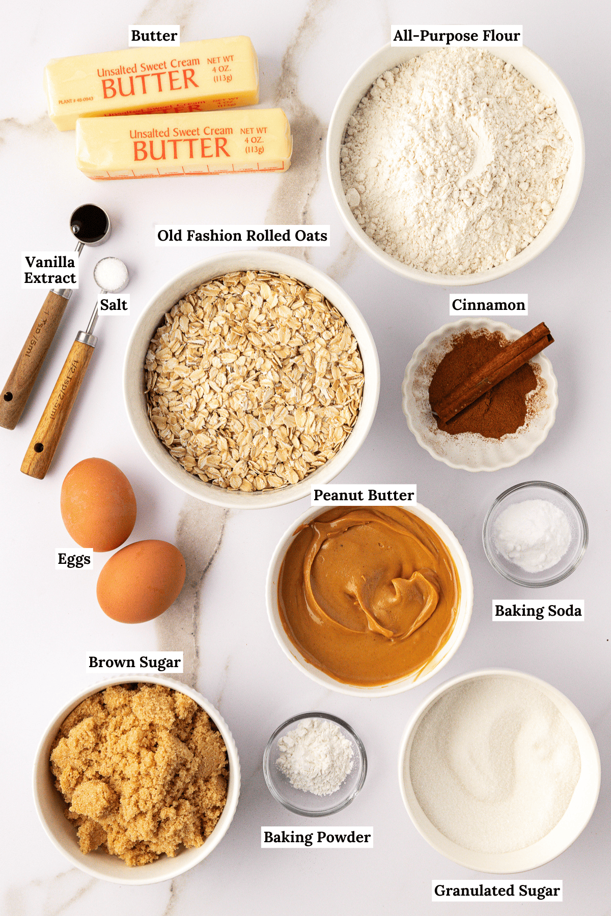 over head view of the ingredients for peanut butter oatmeal cookies including butter, all-purpose flour, vanilla extract, salt, old fashion rolled oats, cinnamon, eggs, peanut butter, baking soda, brown sugar, baking powder and granulated sugar