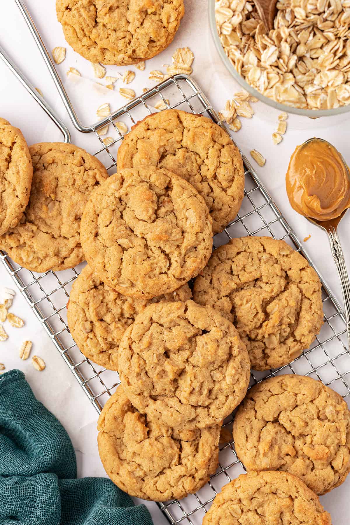 over head view of peanut butter oatmeal cookies scattered on a wire rack, surrounded by a dark teal cloth, scattered oats, a spoonful of peanut butter and bowl of oats