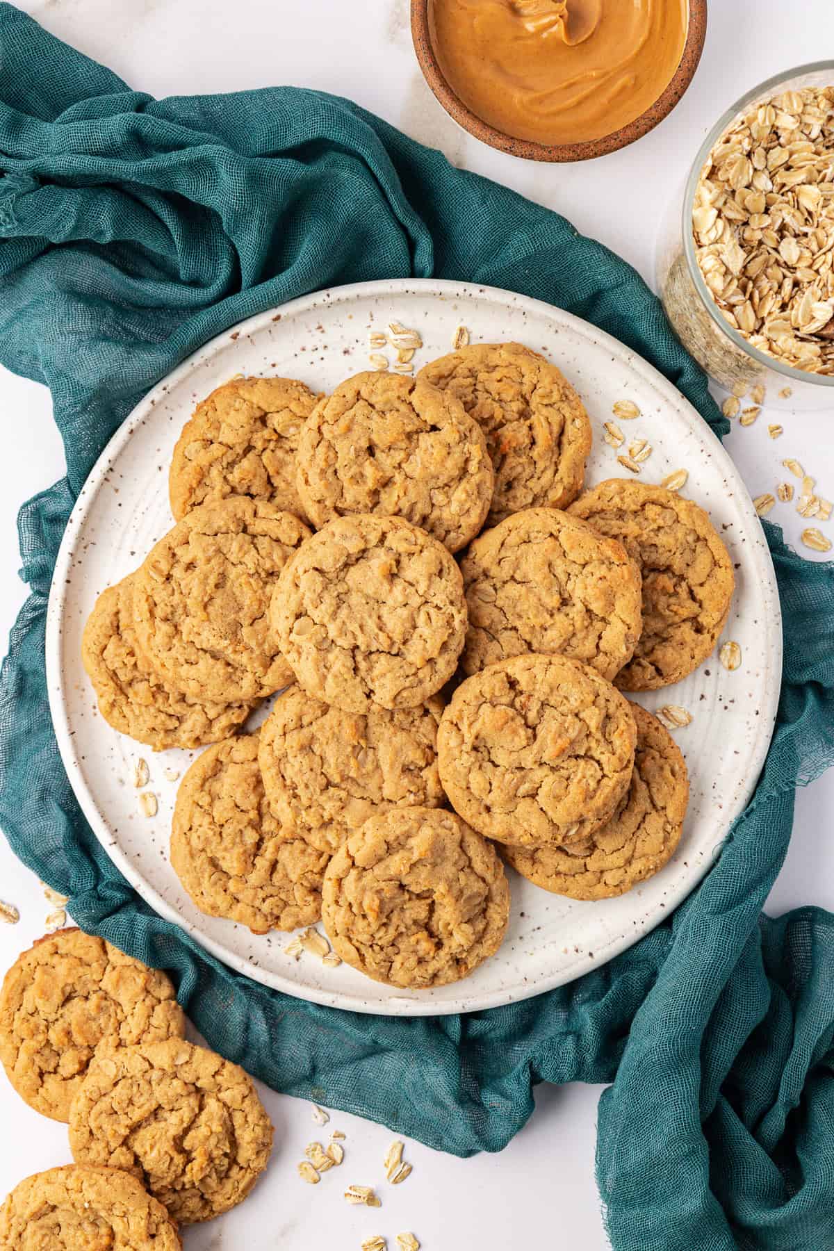 over head view of a plate stacked with peanut butter oatmeal cookies on top of a dark teal cloth, surrounded by more cookies, oats, a small bowl of peanut butter and a glass container of oats