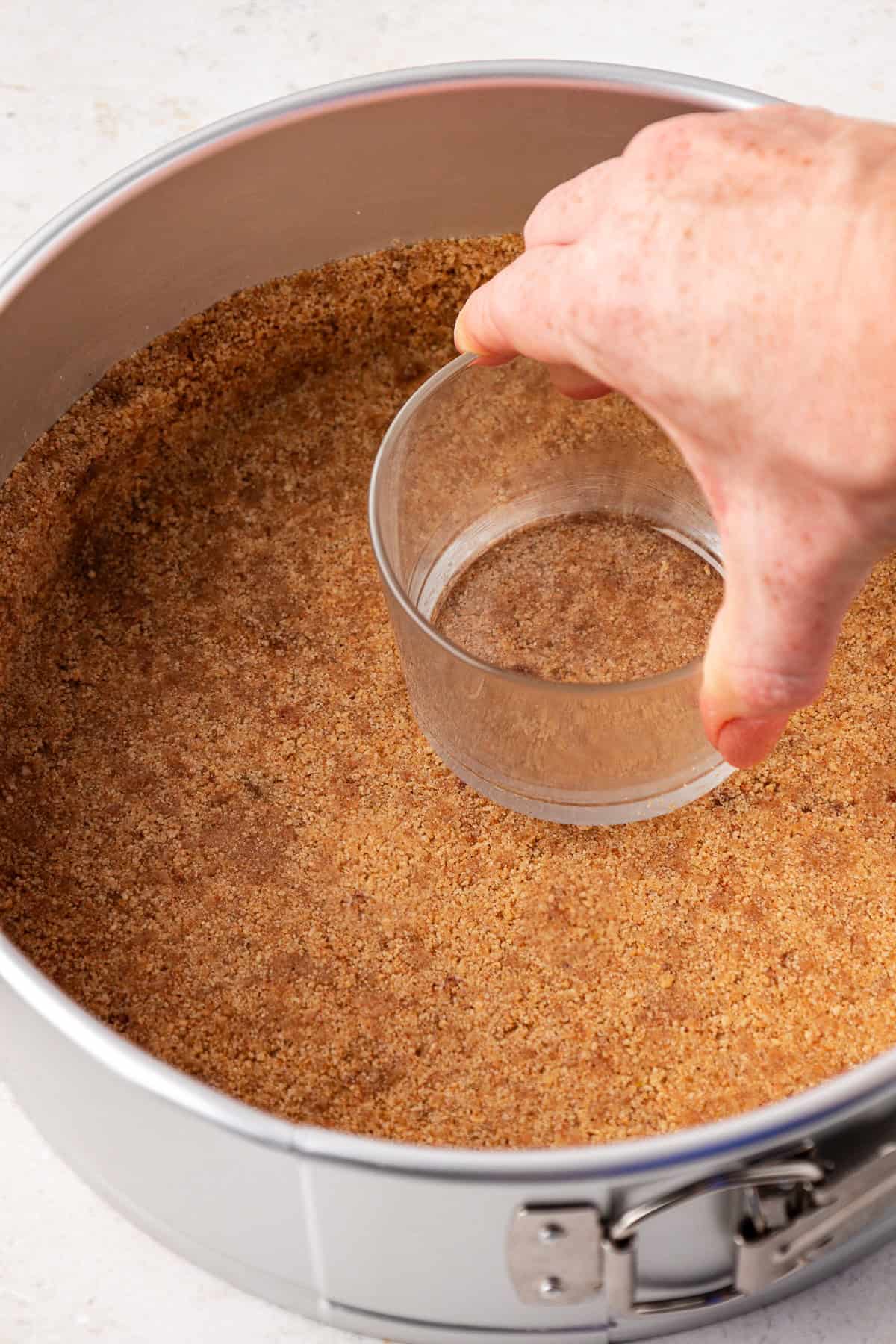 graham cracker crust being pressed down into a springform pan with glass