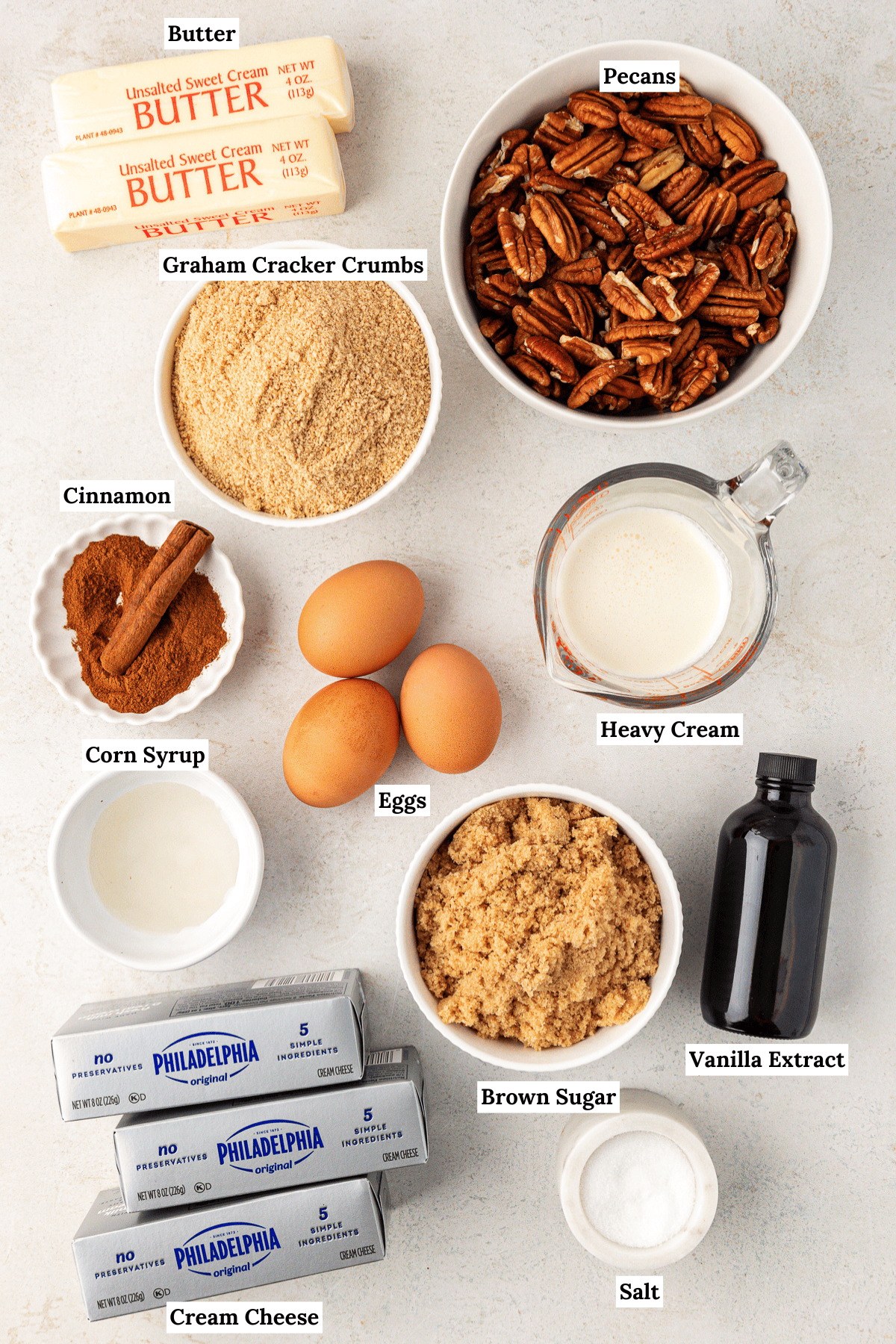 over head view of the ingredients for pecan pie cheesecake including butter, pecans, graham cracker crumbs, cinnamon, corn syrup, eggs, heavy cream, brown sugar, vanilla extract, cream cheese, and salt