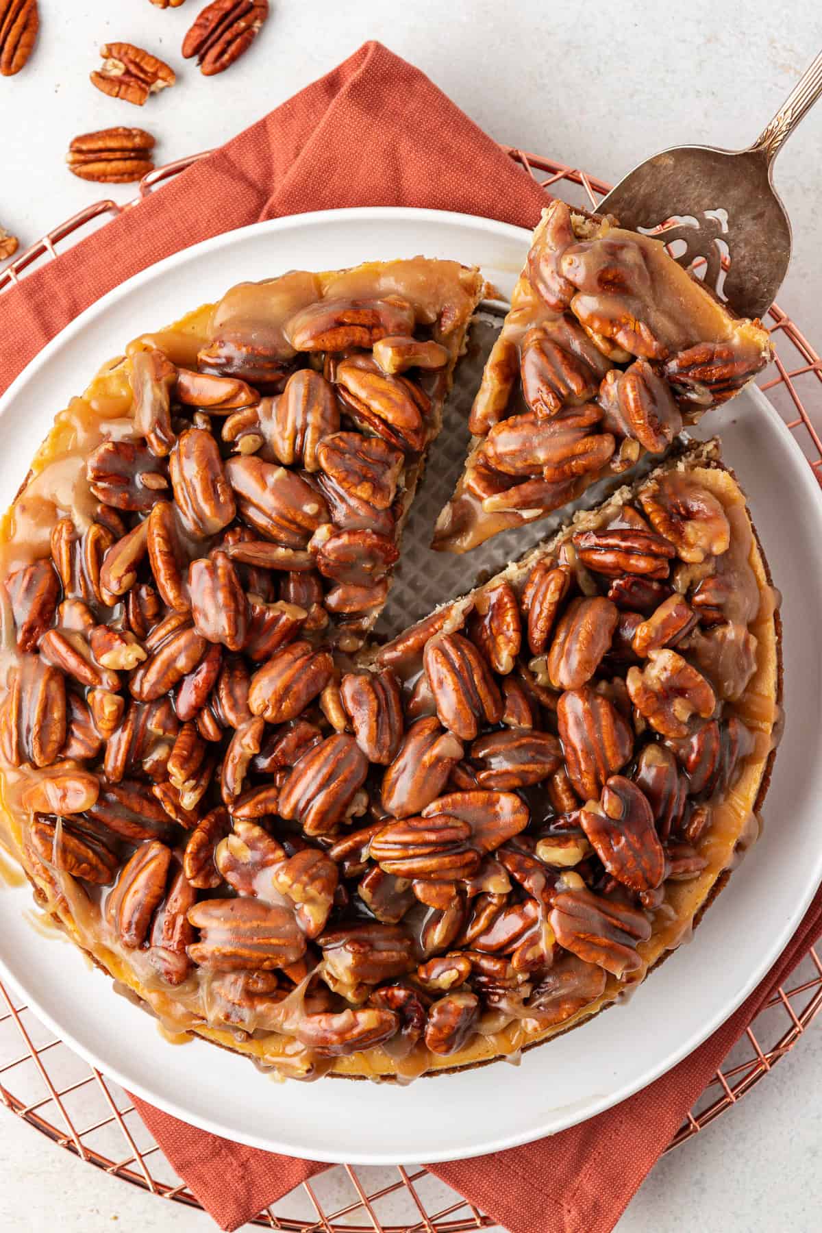 over head view of a pecan pie cheesecake on top of a burnt orange cloth on a wire rack, with one slice being removed by a serving spatula and pecans scattered around