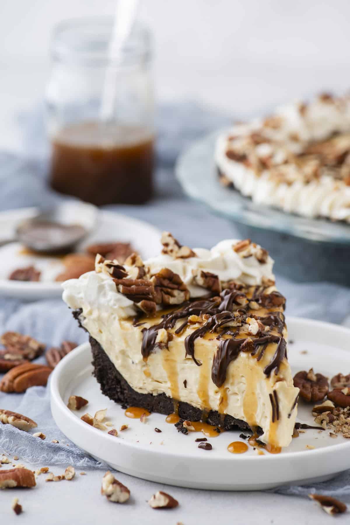 a slice of turtle pie topped with pecans, chocolate and caramel drizzles on a small white plate with pecan pieces scattered around