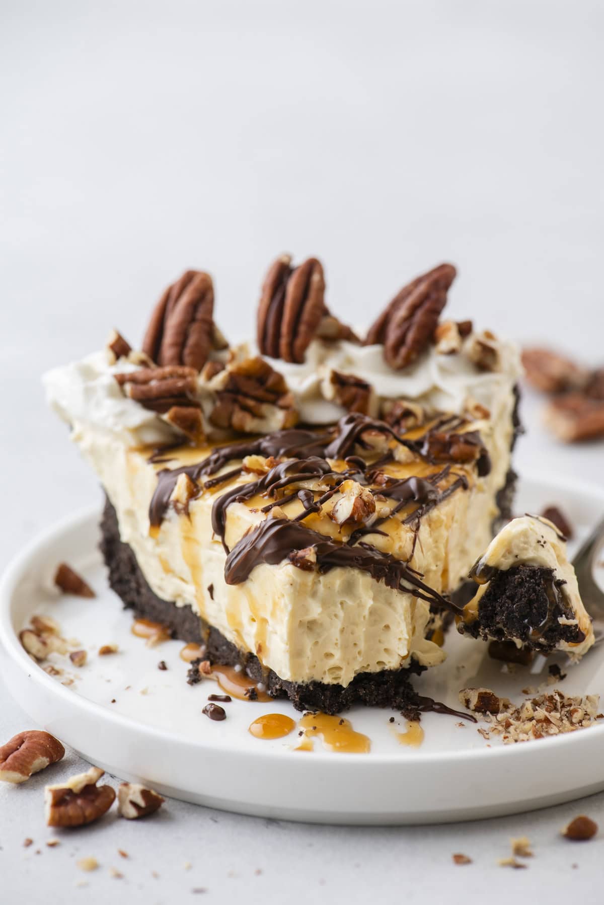 a slice of turtle pie topped with pecans, chocolate and caramel drizzles on a small white plate with pecan pieces scattered around