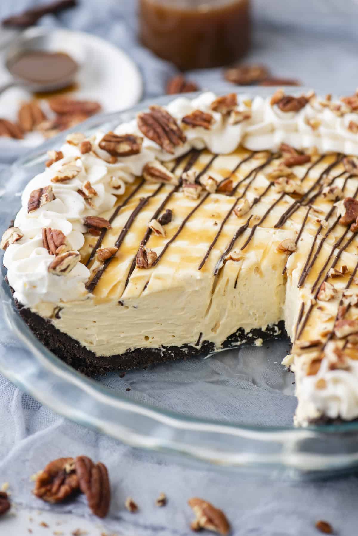 turtle pie in a glass pie dish topped with caramel, chocolate, whip cream and pecans with pecans scattered around