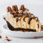 a slice of turtle pie topped with pecans, chocolate and caramel drizzles on a small white plate with pecan pieces scattered around