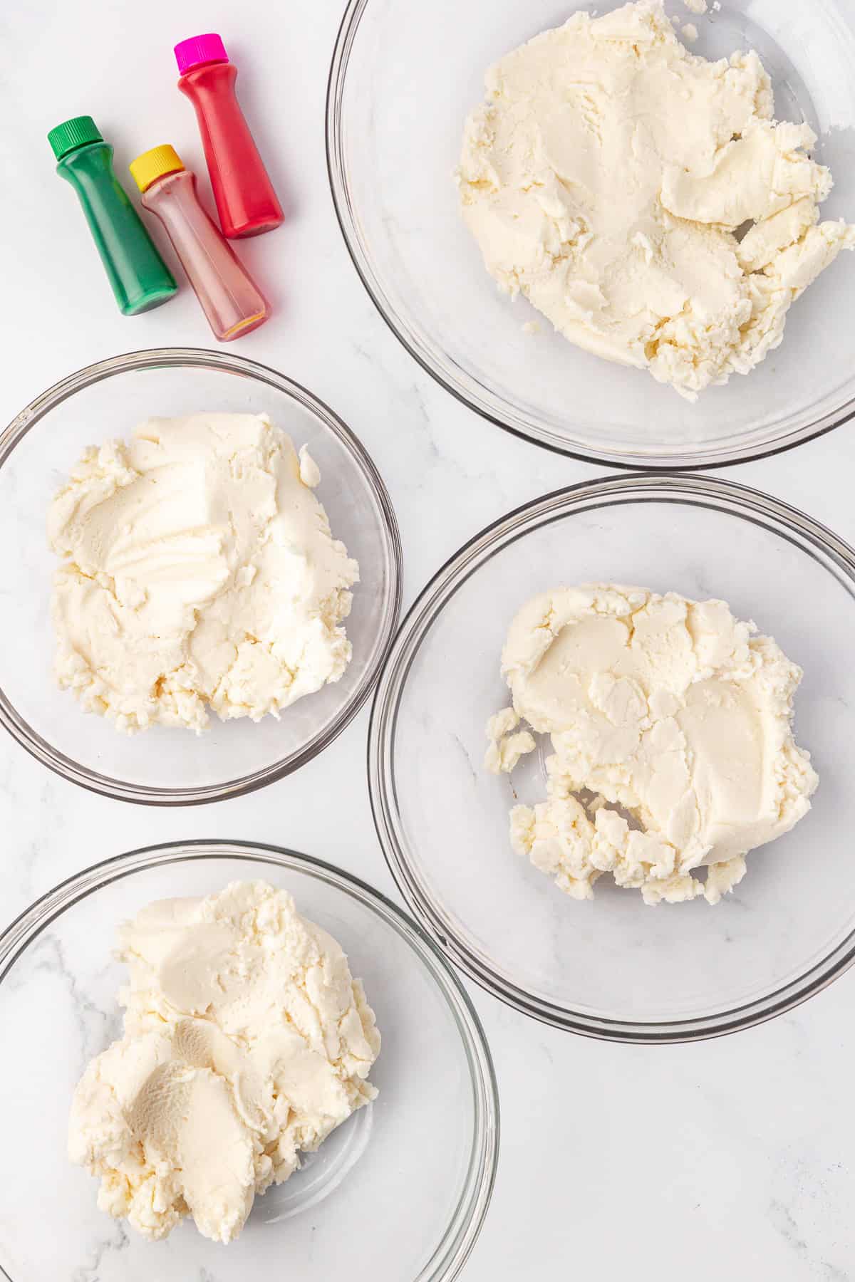 three bottles of food coloring (green, yellow, pink) beside four bowls of white butter mint dough