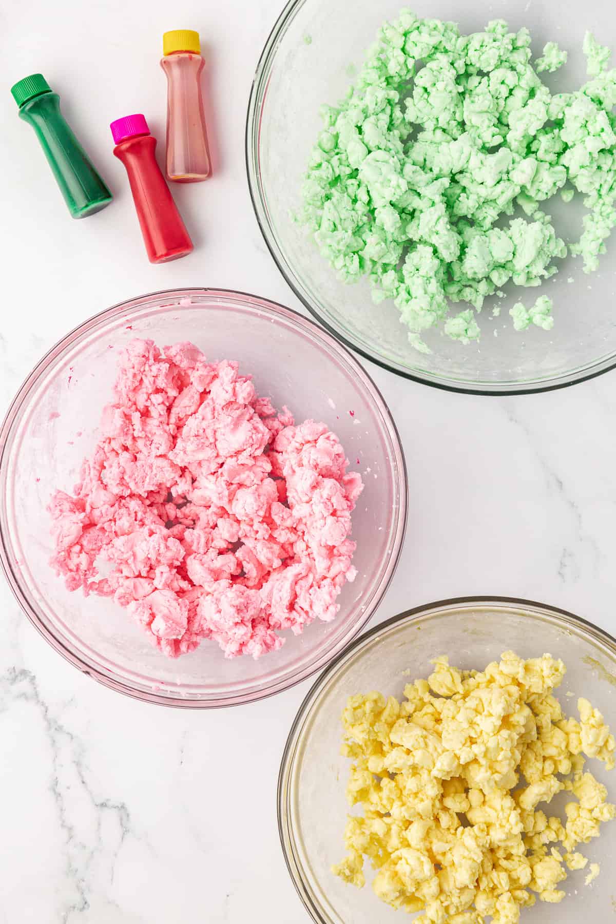 over head view of three glass bowls with butter mint dough and three different colors of food coloring: green, pink, and yellow