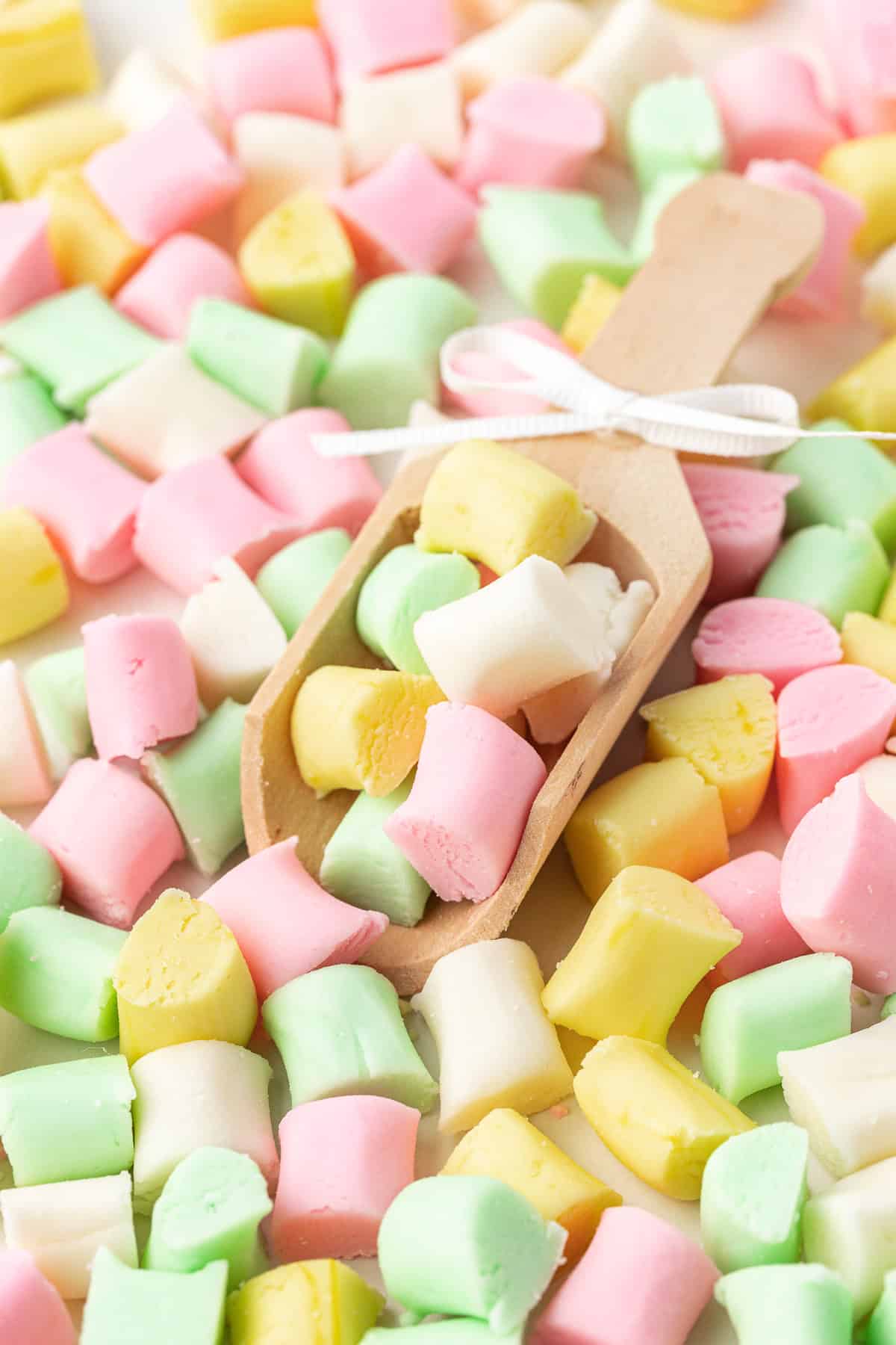 a pile of pink, white, green and yellow butter mints with one small wood scoop with a white bow on it full of mints in the middle