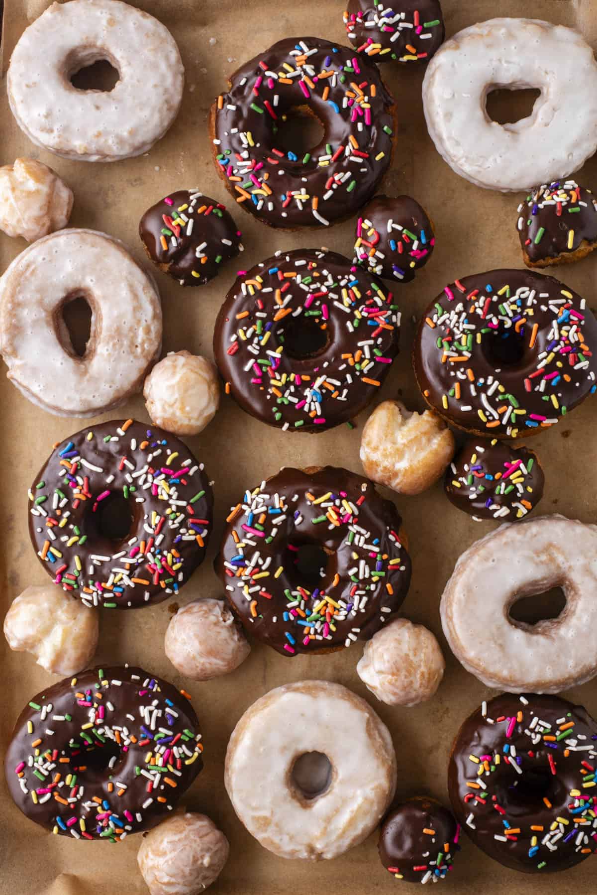 over head view of scattered cake donuts, a mix of donut holes, regular glazed donuts and chocolate glazed with sprinkles