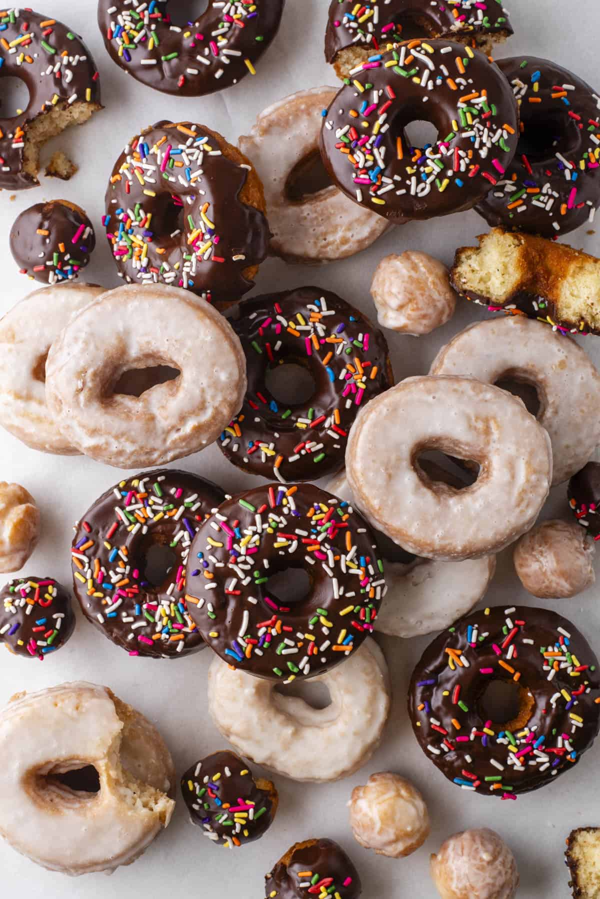 over head view of scattered and stacked cake donuts, a mix of donut holes, regular glazed donuts and chocolate glazed with sprinkles