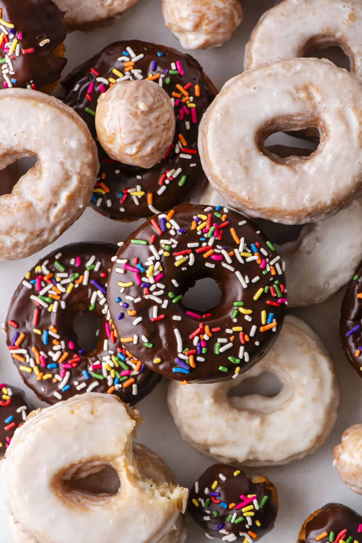 over head view of scattered and stacked cake donuts, a mix of donut holes, regular glazed donuts and chocolate glazed with sprinkles