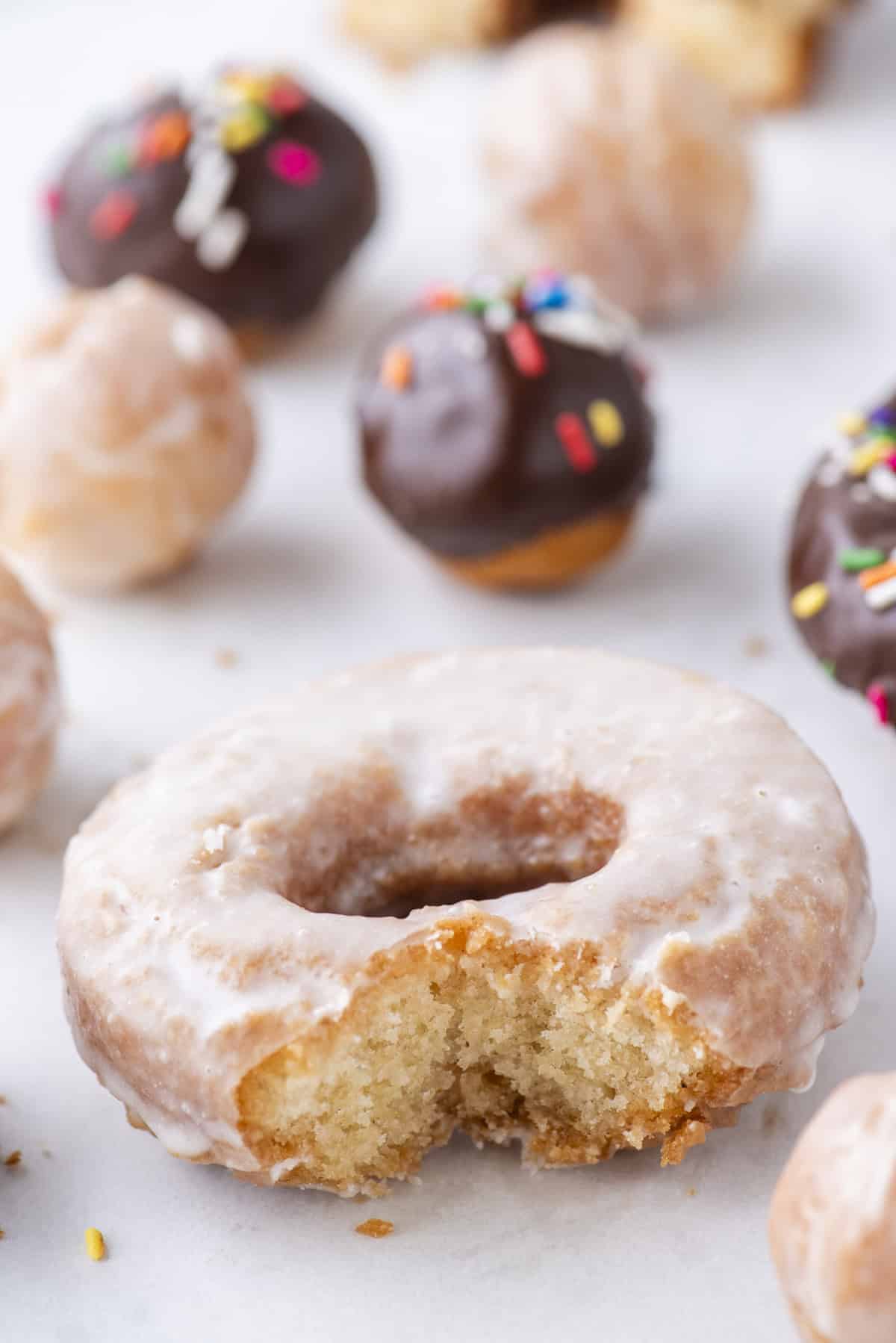 a glazed cake donut with a bite missing out, surrounded by donut holes (a mix of glazed and chocolate glazed with sprinkles)