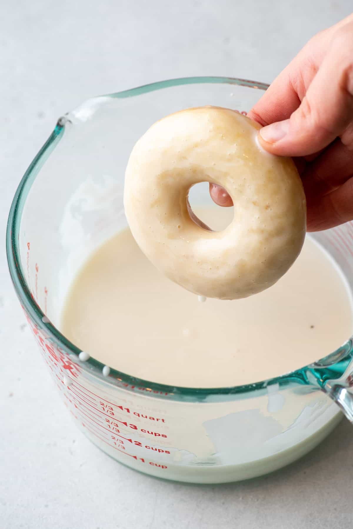 a cake donut over top of a large measuring cup of glaze, being held by a hand with glaze dripping off of it