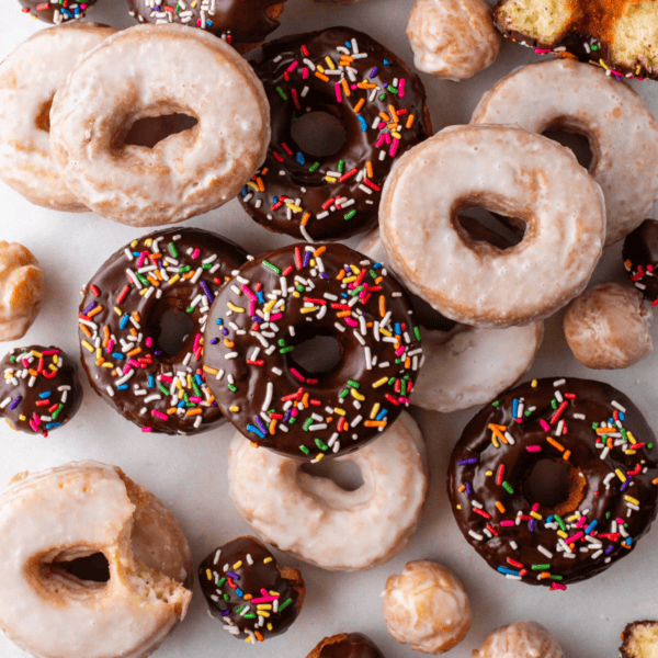 over head view of scattered and stacked cake donuts, a mix of donut holes, regular glazed donuts and chocolate glazed with sprinkles