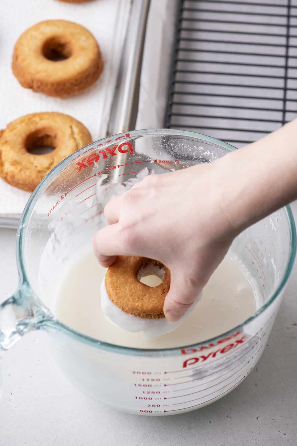 a donut being dipped into a large measuring cup full of glaze beside a wire rack and a baking sheet with donuts on it