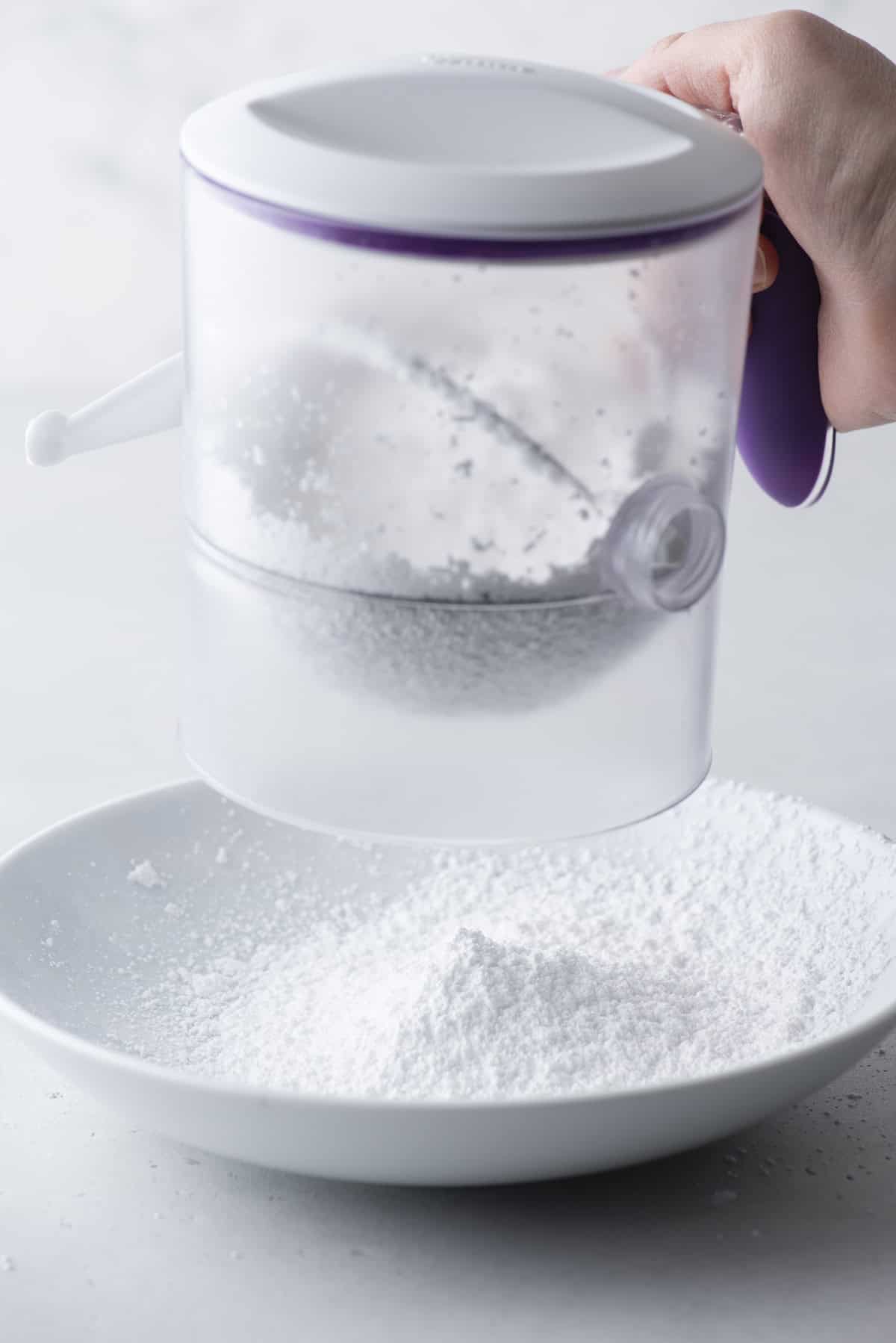 a sifter being held over a white bowl, sifting powdered sugar into the bowl