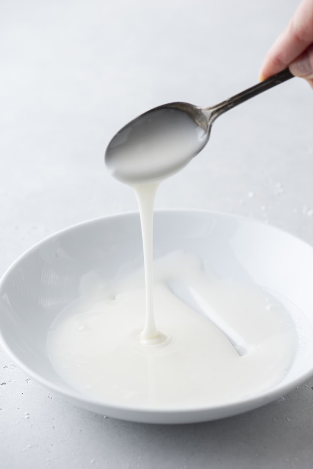 donut glaze in a white bowl with a spoon being held above it drizzling glaze down into the bowl