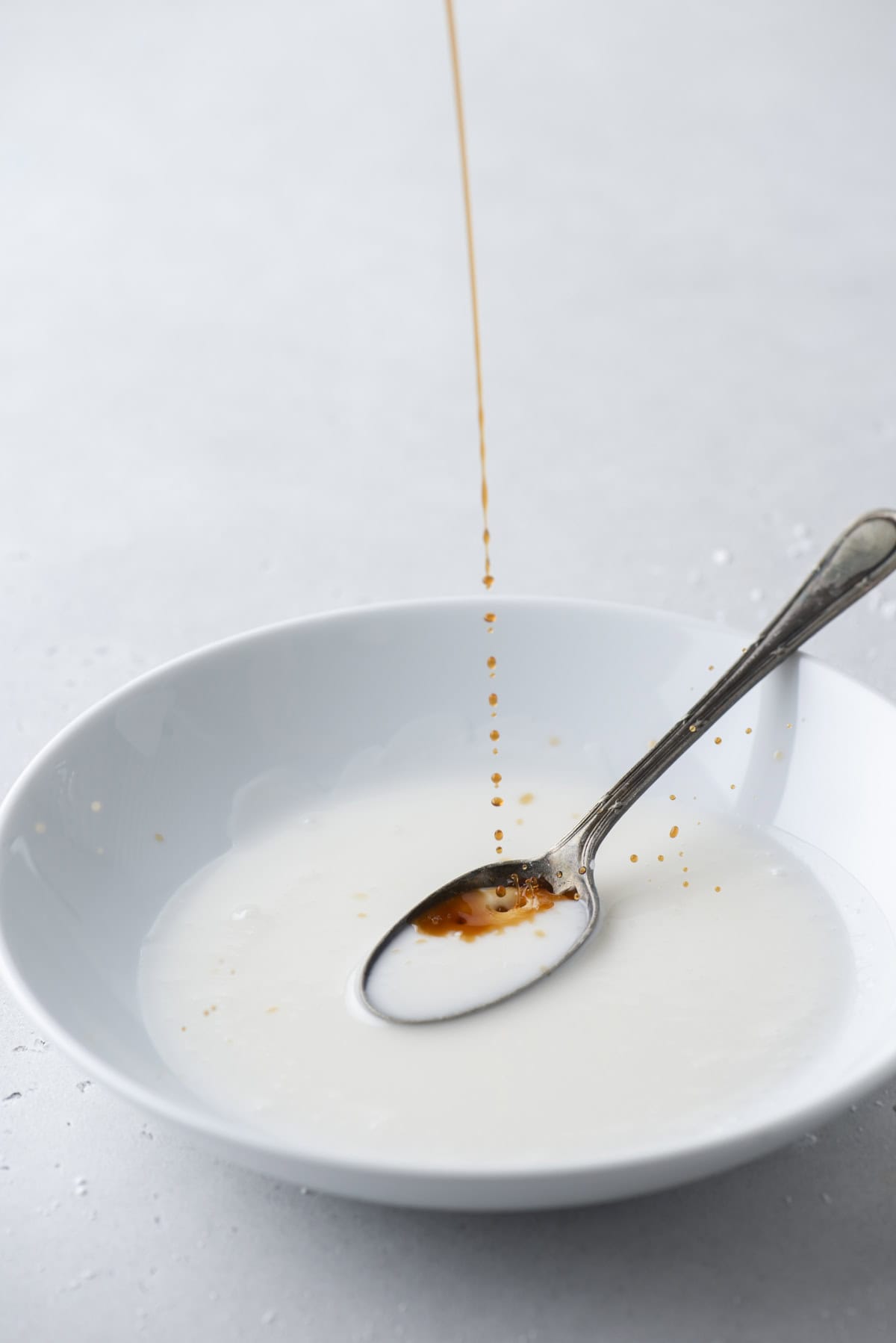 vanilla extract being poured into a white bowl of donut glaze with a spoon in it