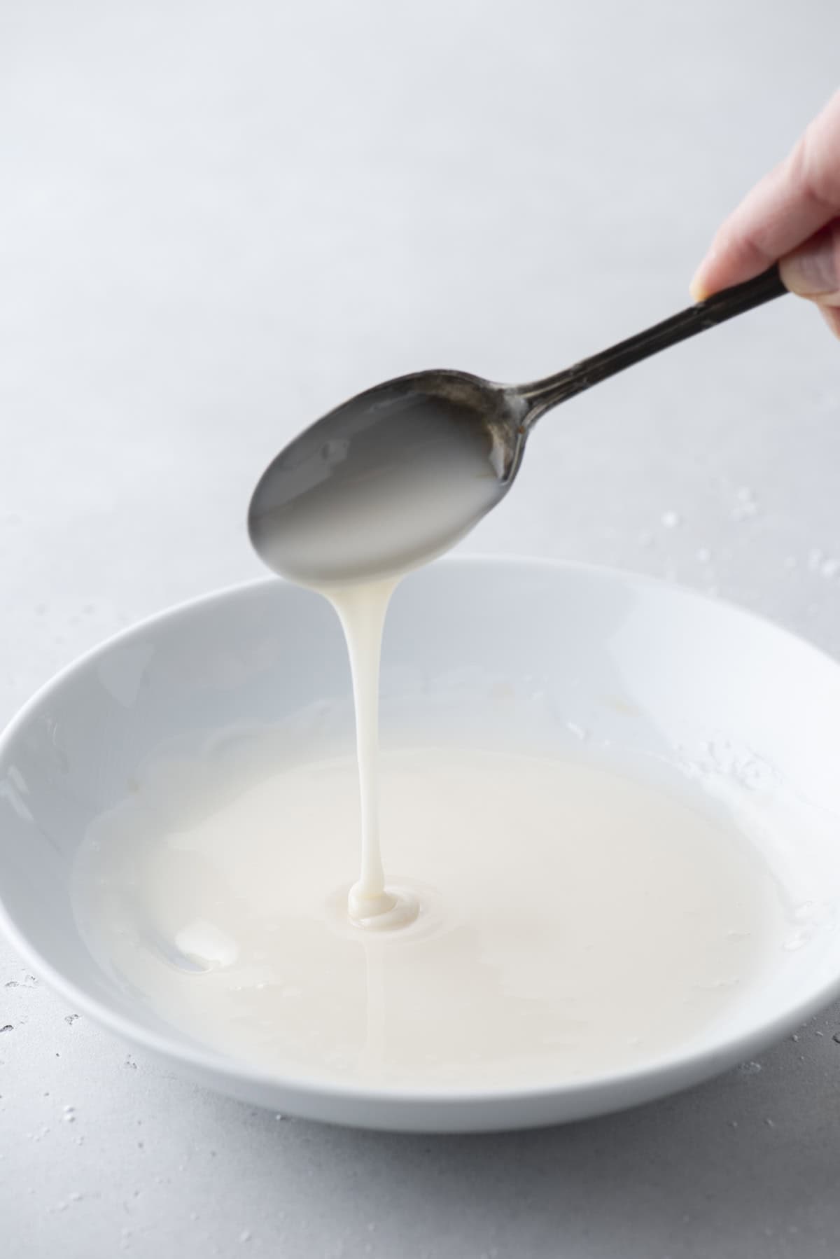 donut glaze in a white bowl with a spoon being held above it drizzling glaze down into the bowl