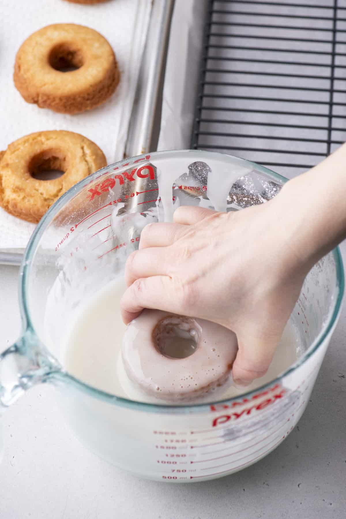 a donut being dipped into a large measuring cup full of glaze beside a wire rack and a baking sheet with donuts on it