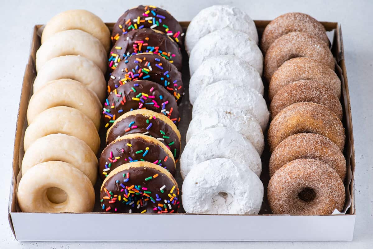 a box filled with rows of donuts with one row of glazed, then chocolate sprinkle, powdered and cinnamon sugar