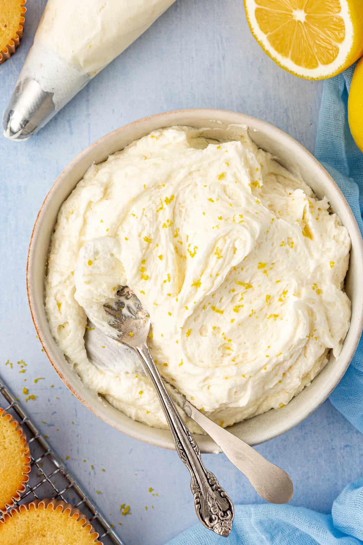 a bowl of lemon frosting topped with lemon zest with spoons in it surrounded by a piping bag of frosting, cupcakes and fresh lemons