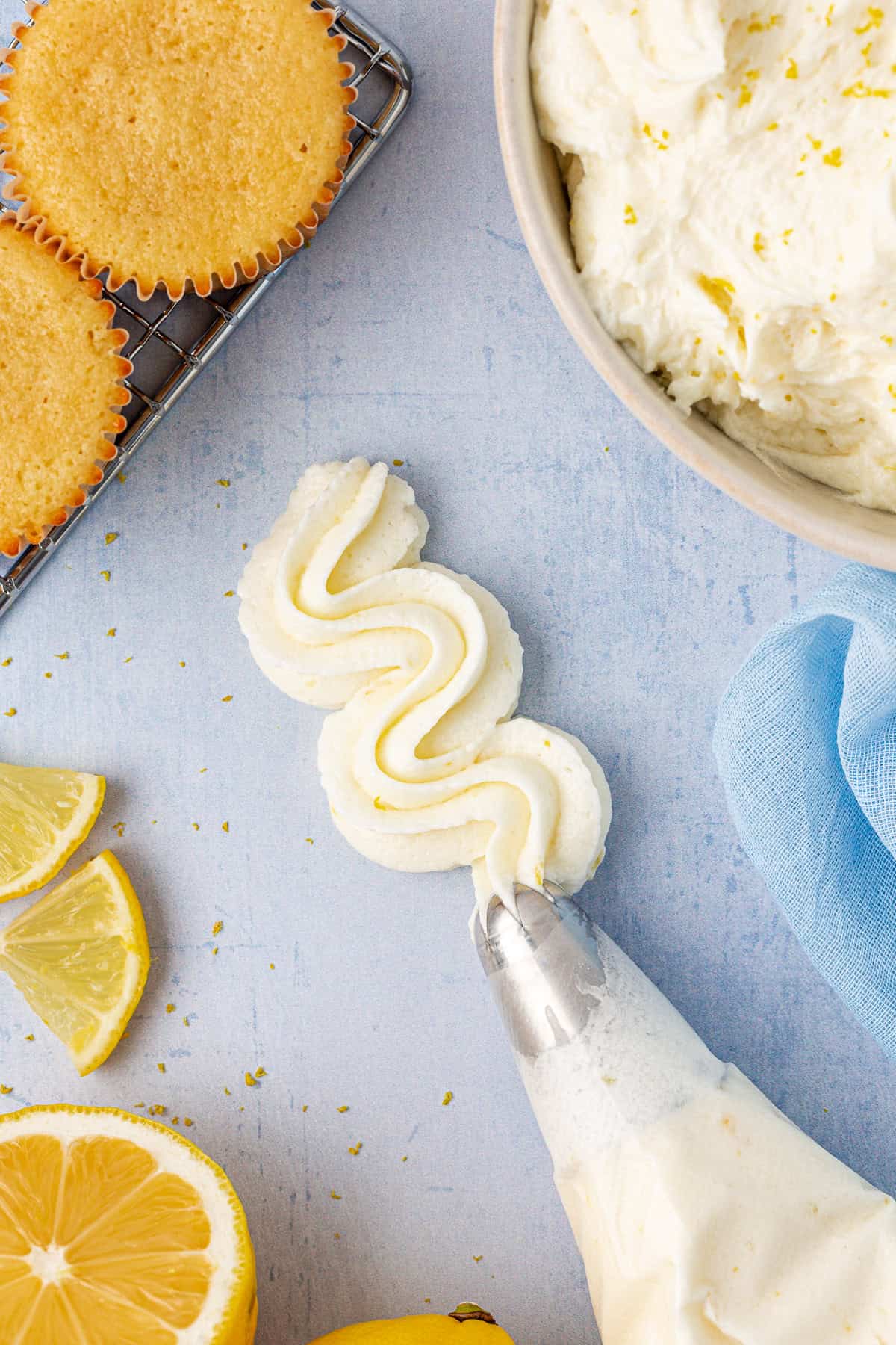 a piping bag spreading lemon frosting in the middle of fresh lemon slices, cupcakes on a wire rack, and a bowl of frosting