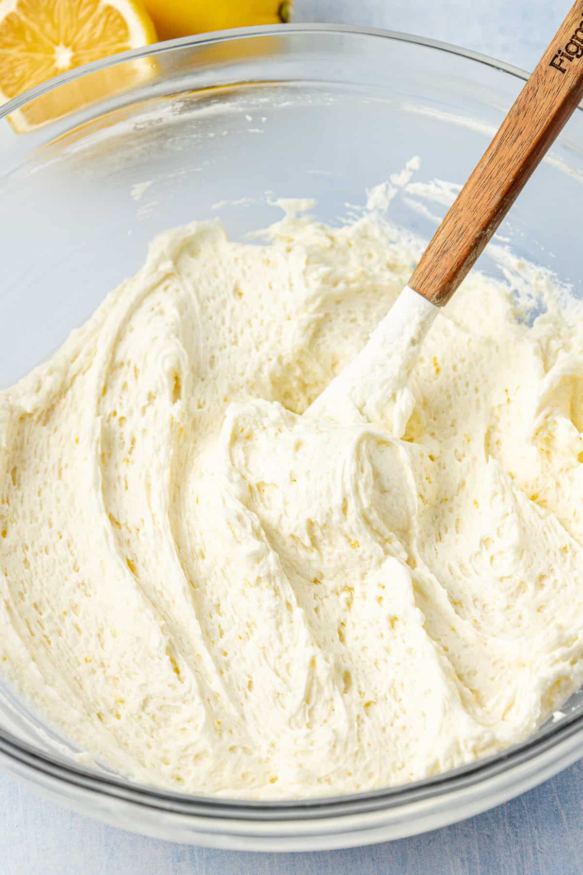 frosting in a clear glass bowl with a wooden spoon in it and fresh lemons in the background