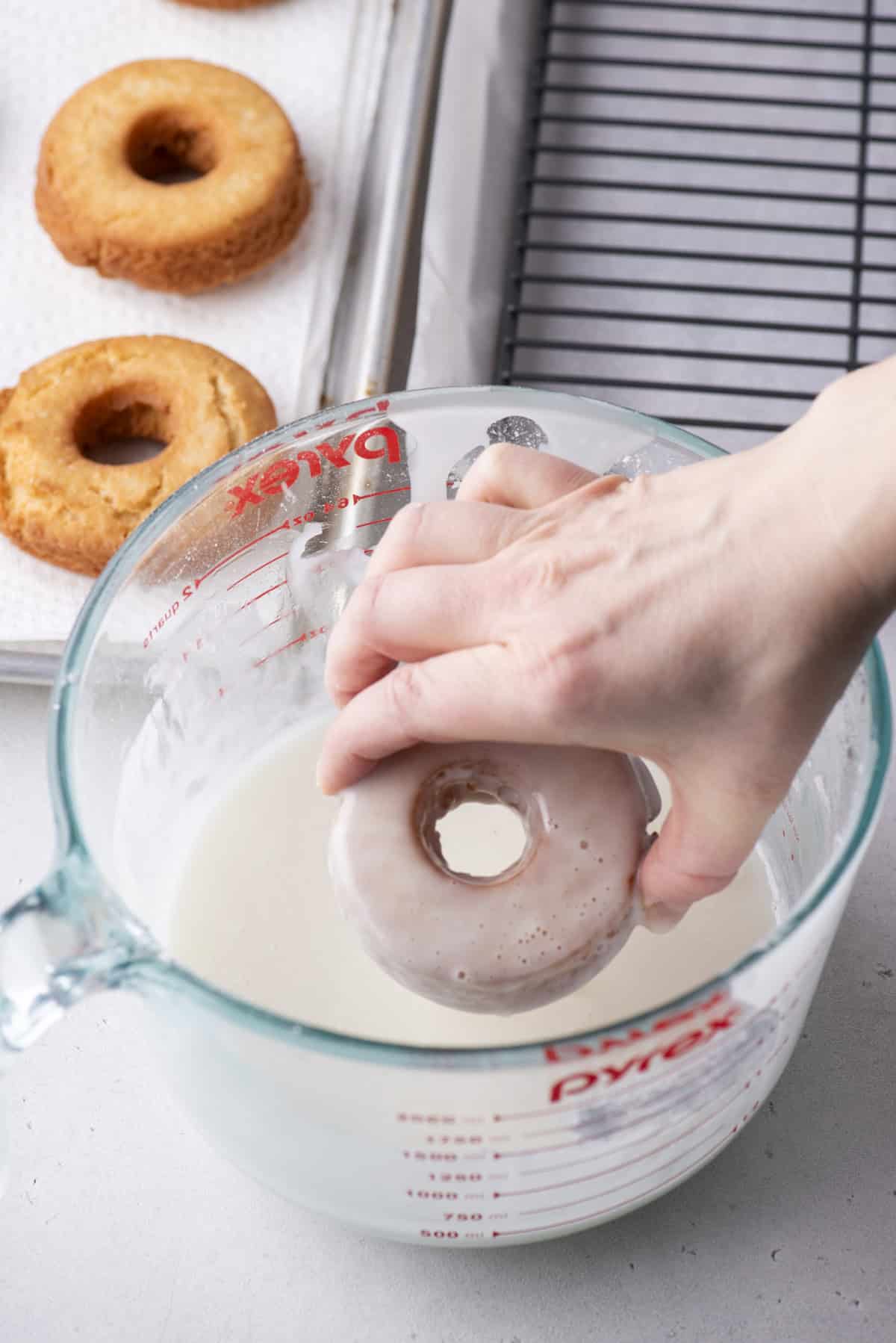 a donut being dipped by hand into a large measuring cup full of donut glaze, beside a baking sheet with more donuts and a wire rack