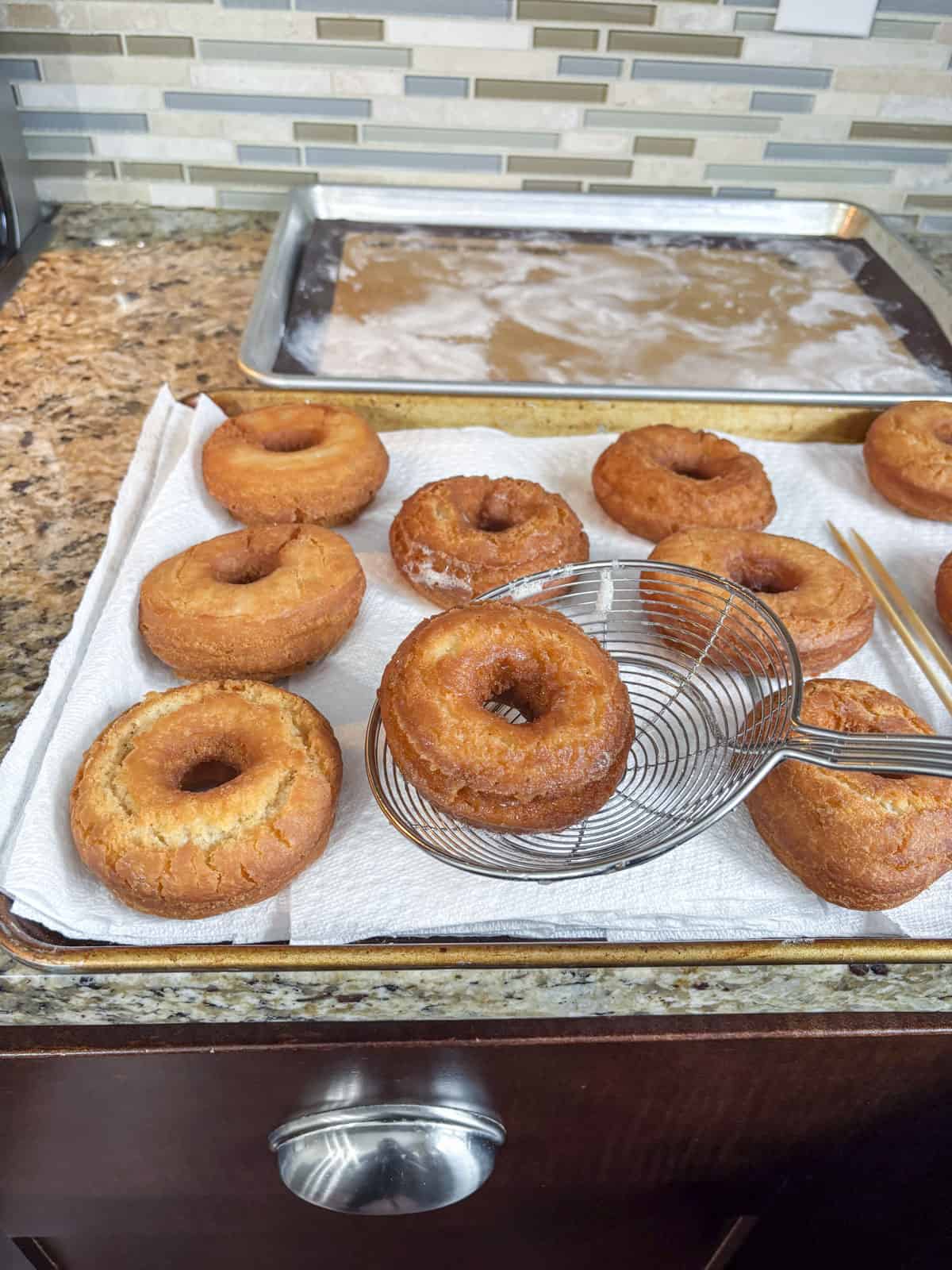 a donut being placed with a spider strainer onto a baking sheet lined with paper towels with more donuts on it